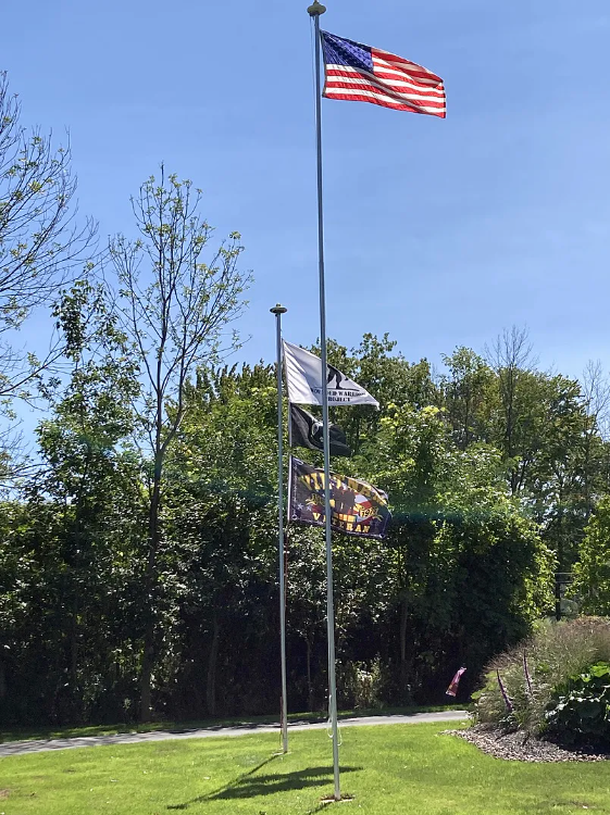 Flagpole with American flag and other flags against a clear blue sky with trees in the background.